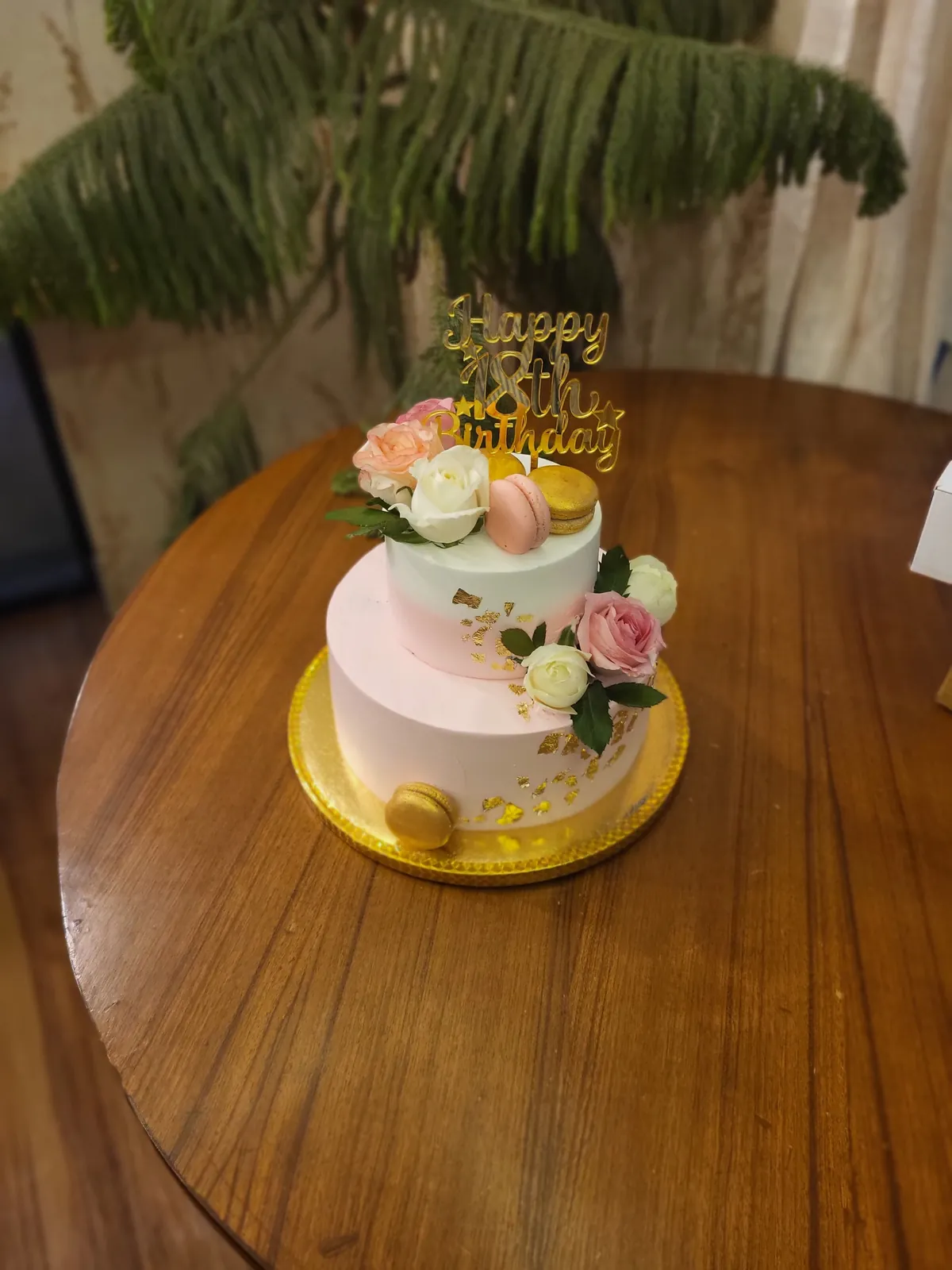 A two-tier birthday cake in pink and cream, decorated with fresh roses, macarons and gold leaf