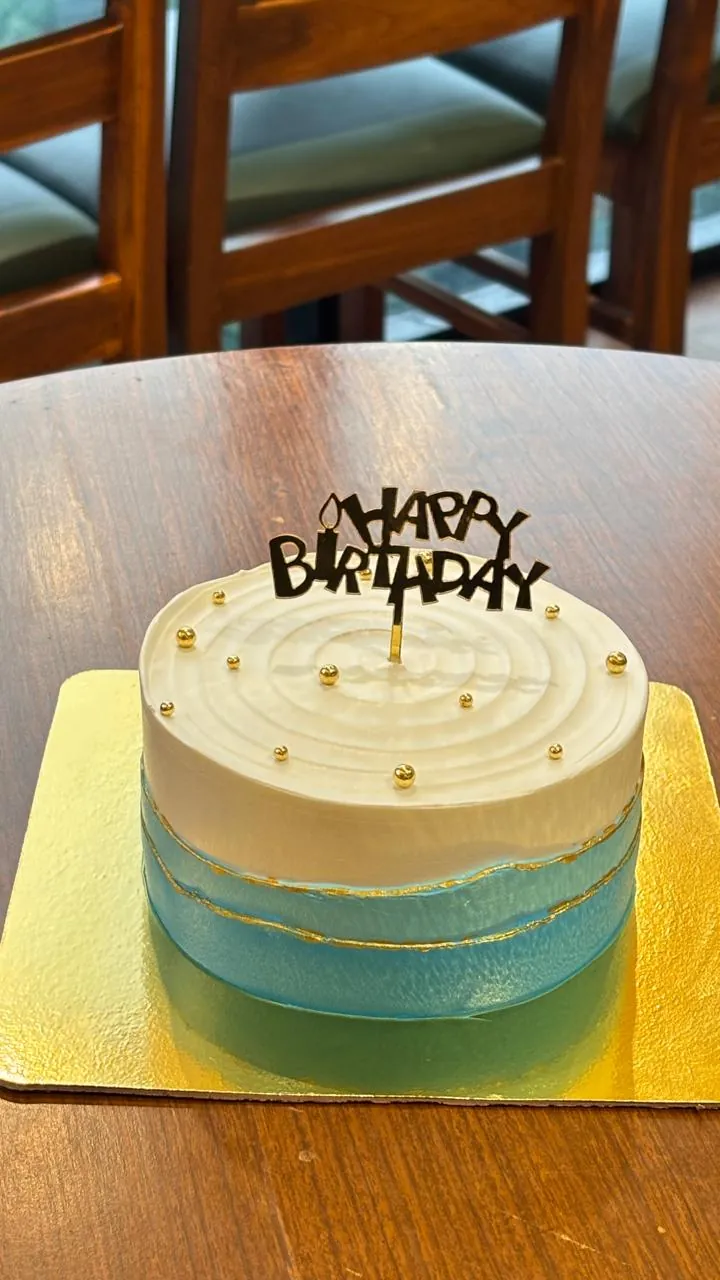 A white and teal ombré cake with gold pearls and a gold birthday topper, on a café table